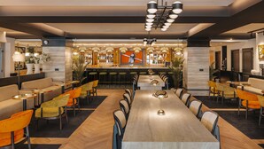 A modern restaurant with a long communal table, multiple bar stools, and a bar area with a variety of bottles and a large screen. - Marty Hotel Bordeaux, Tapestry Collection by Hilton (Bordeaux)