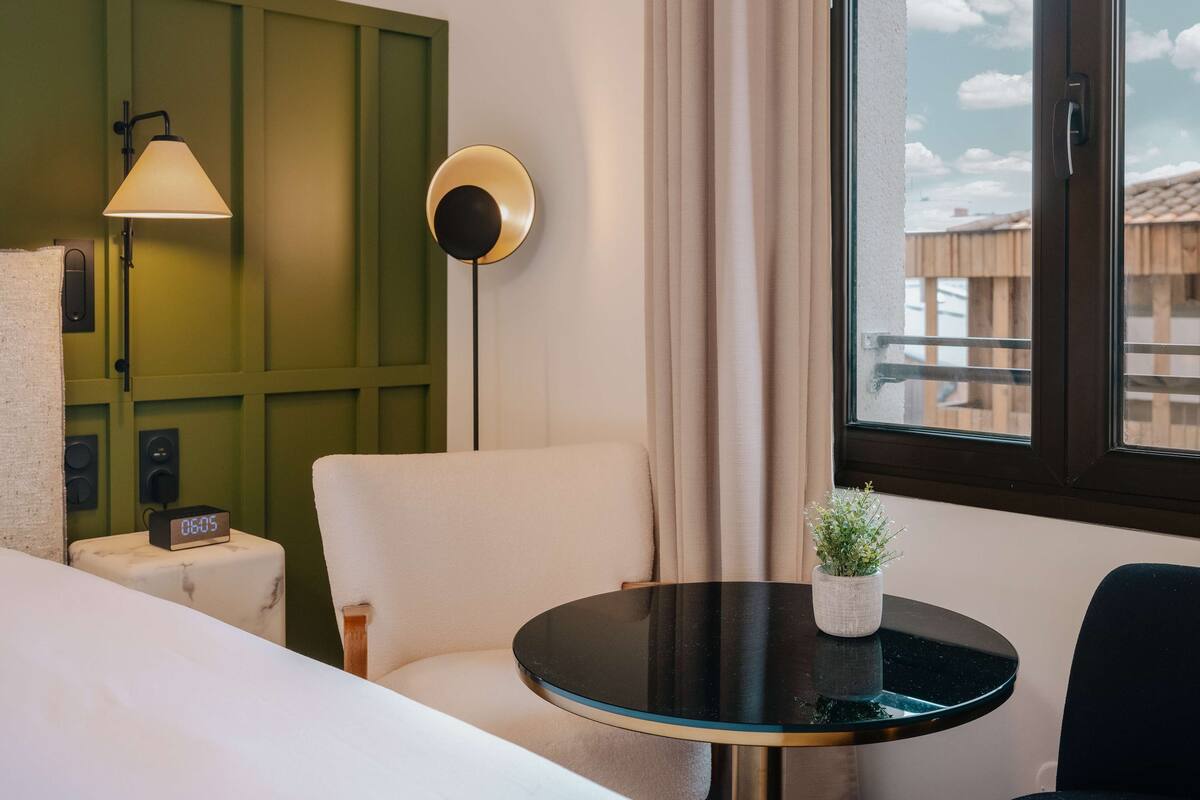 A hotel room with a bed, a round table with a potted plant, a green accent wall, a lamp, and a window with a view of buildings.