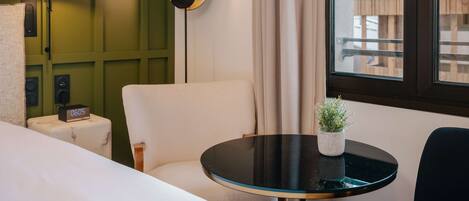 Habitación de hotel con cama, una mesa redonda con planta en maceta, pared de acento verde, lámpara y ventana con vista a edificios.