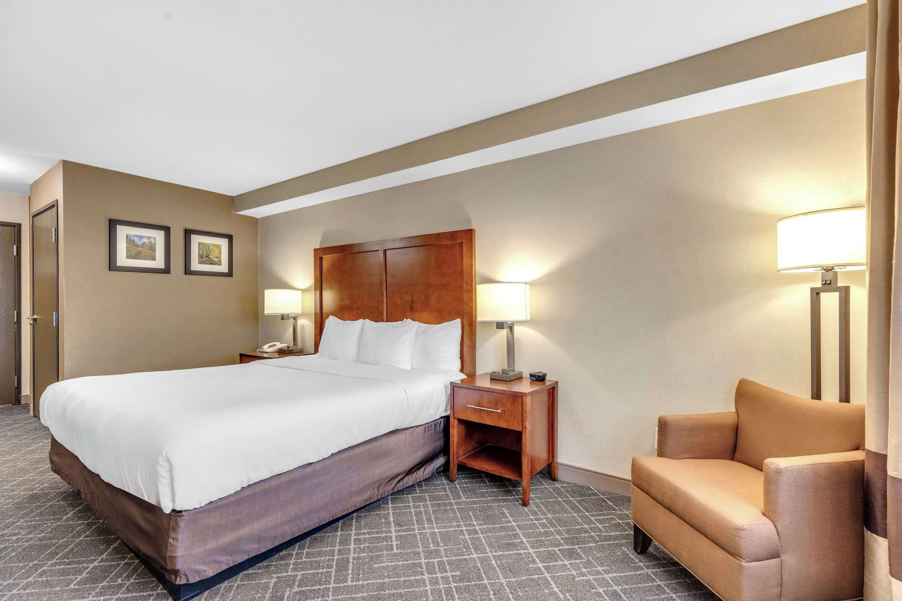 A hotel room with a large bed, a wooden nightstand with a lamp, a chair, and two framed pictures on the wall.
