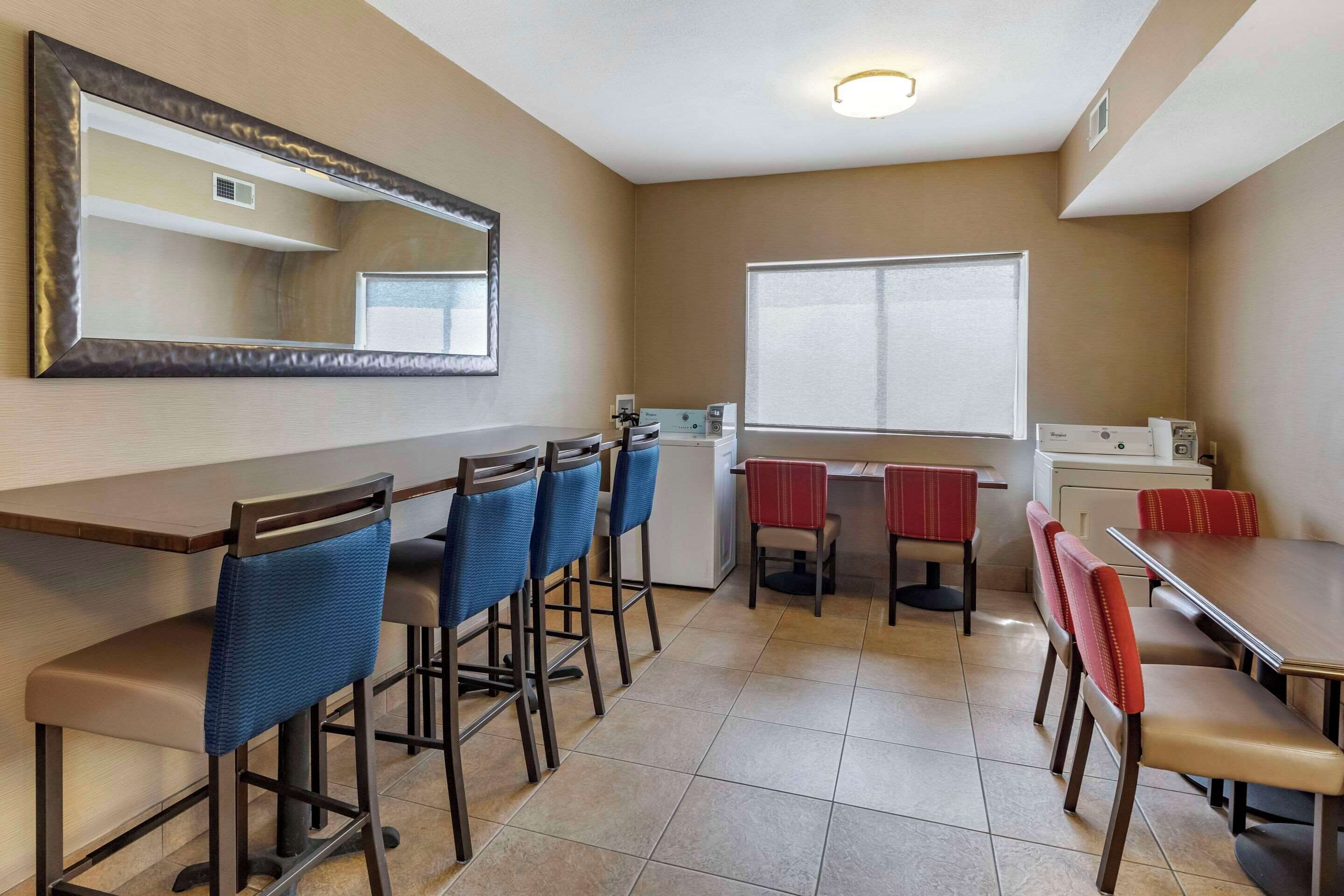 Habitación de hotel con un espejo grande, una ventana con persianas, una kitchenette y un área de comedor con taburetes altos y una mesa.