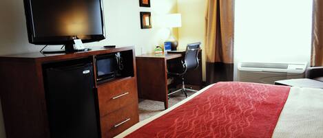 A hotel room with a bed, a desk with a computer, a television, a mini-fridge, and a lamp.