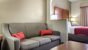 A hotel room with a grey sofa, red pillows, a wooden side table, a bed with a red blanket, and a mirror above the sofa. - Comfort Inn & Suites Covington - Mandeville (Covington)