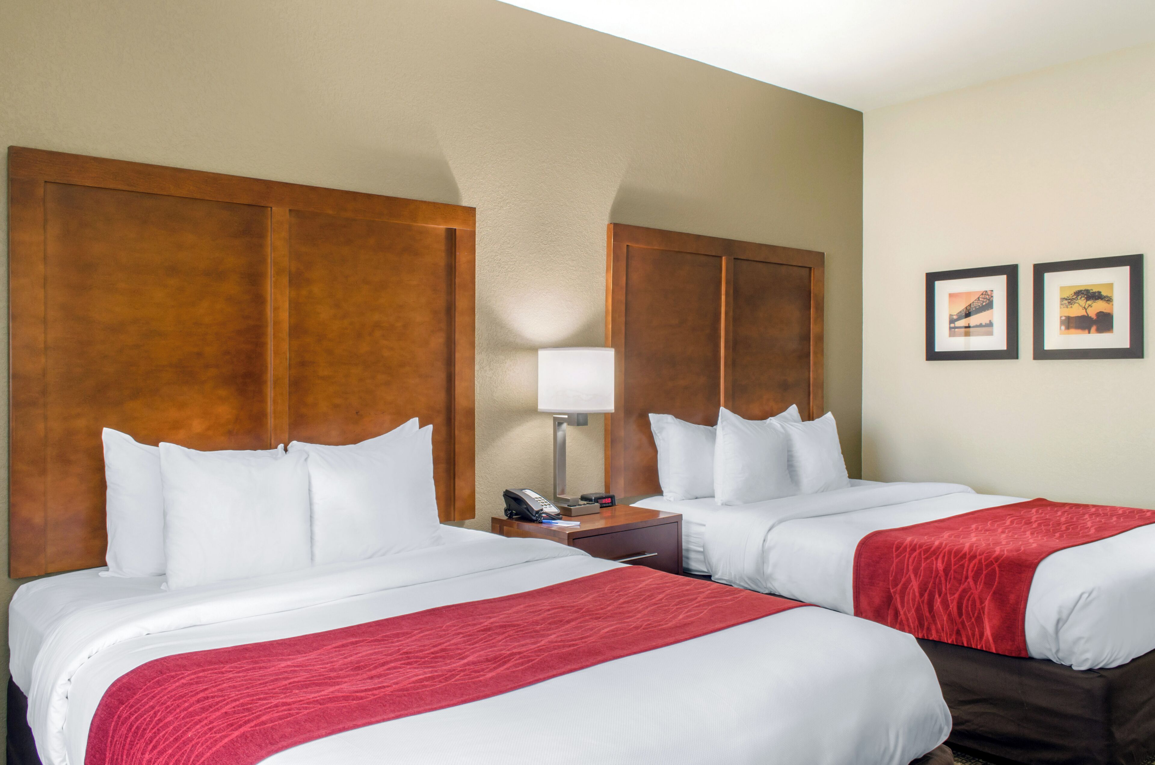 A hotel room with two beds, a wooden headboard, a bedside table with a lamp, and two framed pictures on the wall.