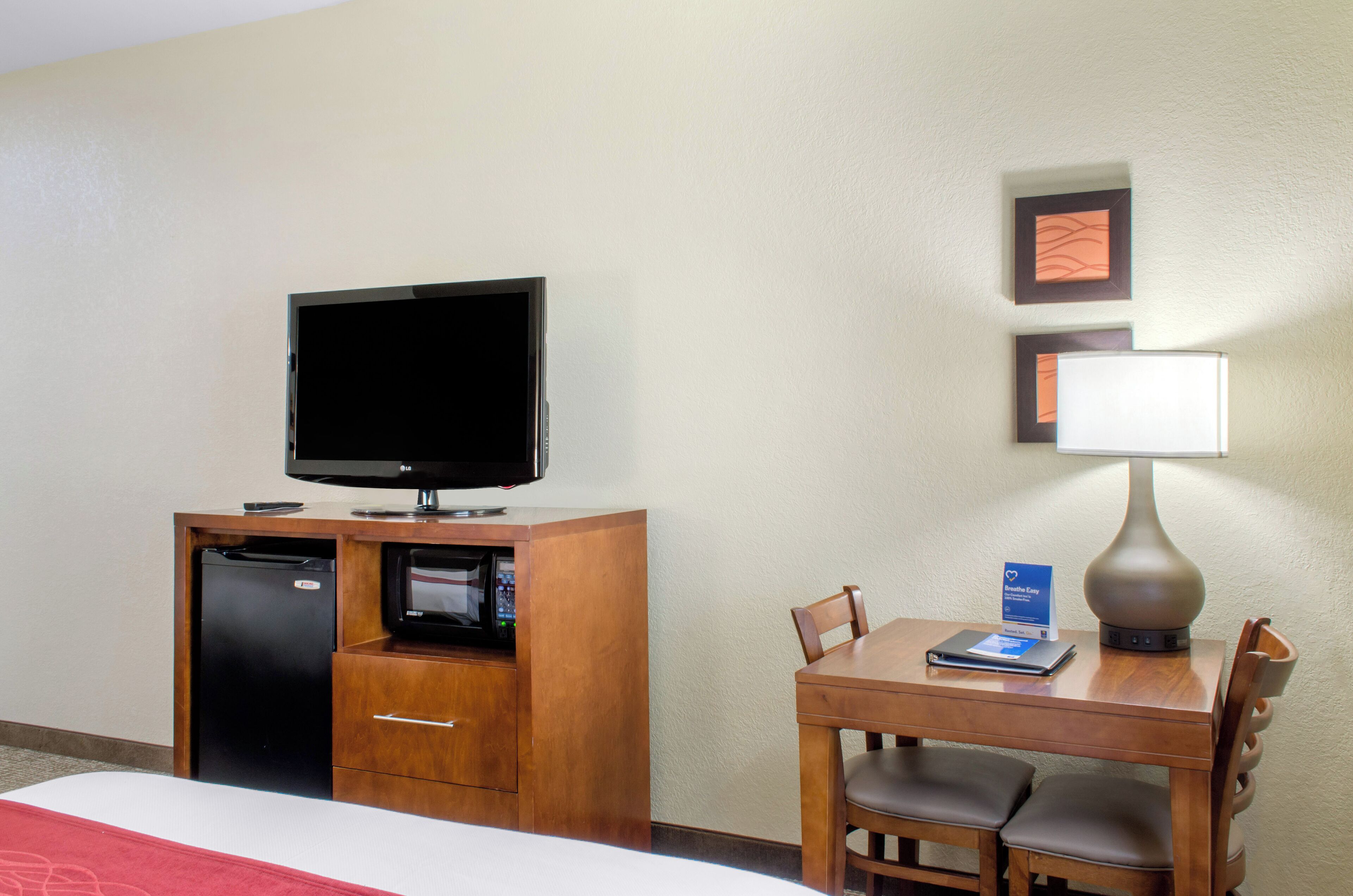 A hotel room with a bed, a desk with a lamp, a television, a refrigerator, and a microwave.