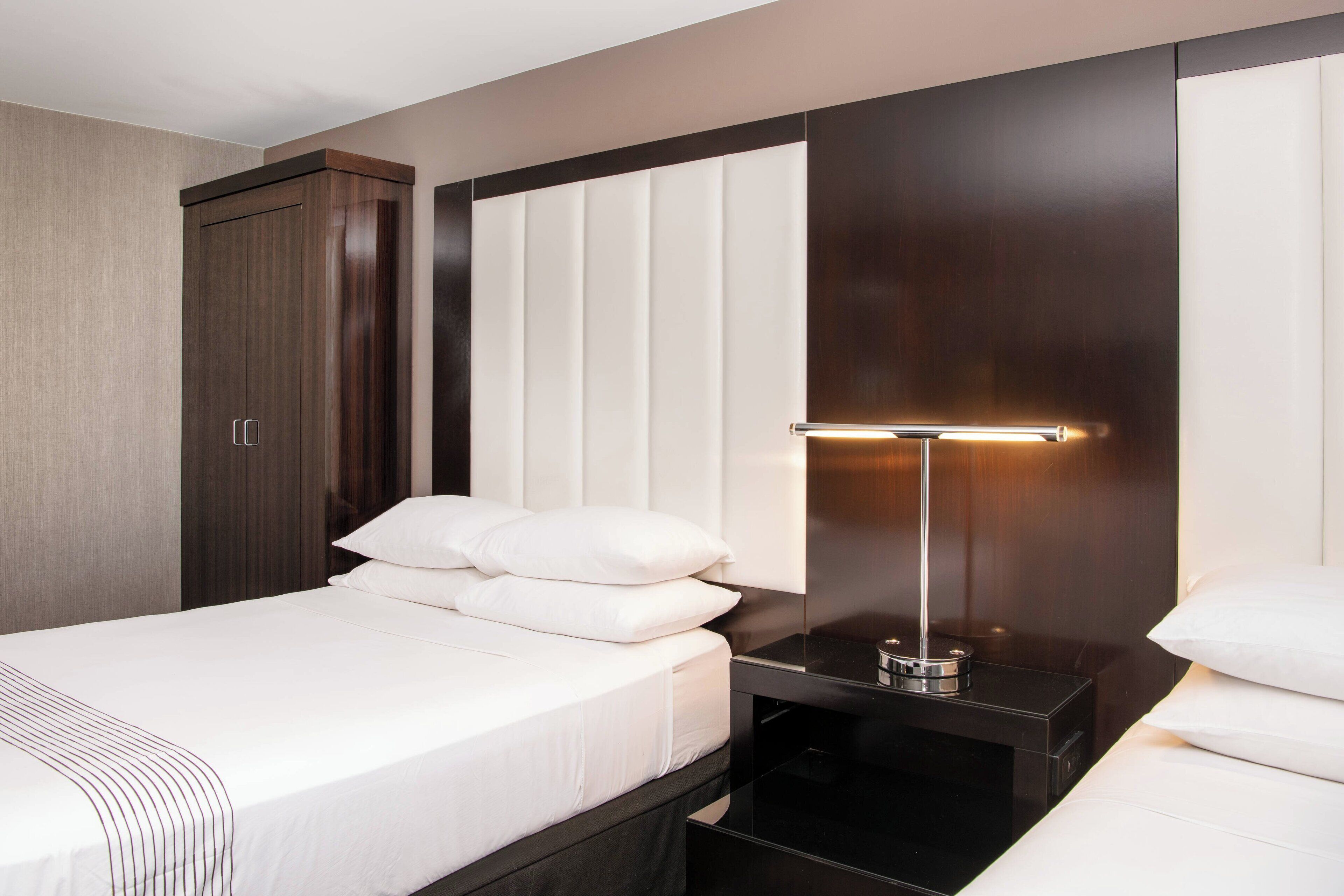 A modern hotel room with a large bed, a bedside table with a lamp, and a wardrobe.