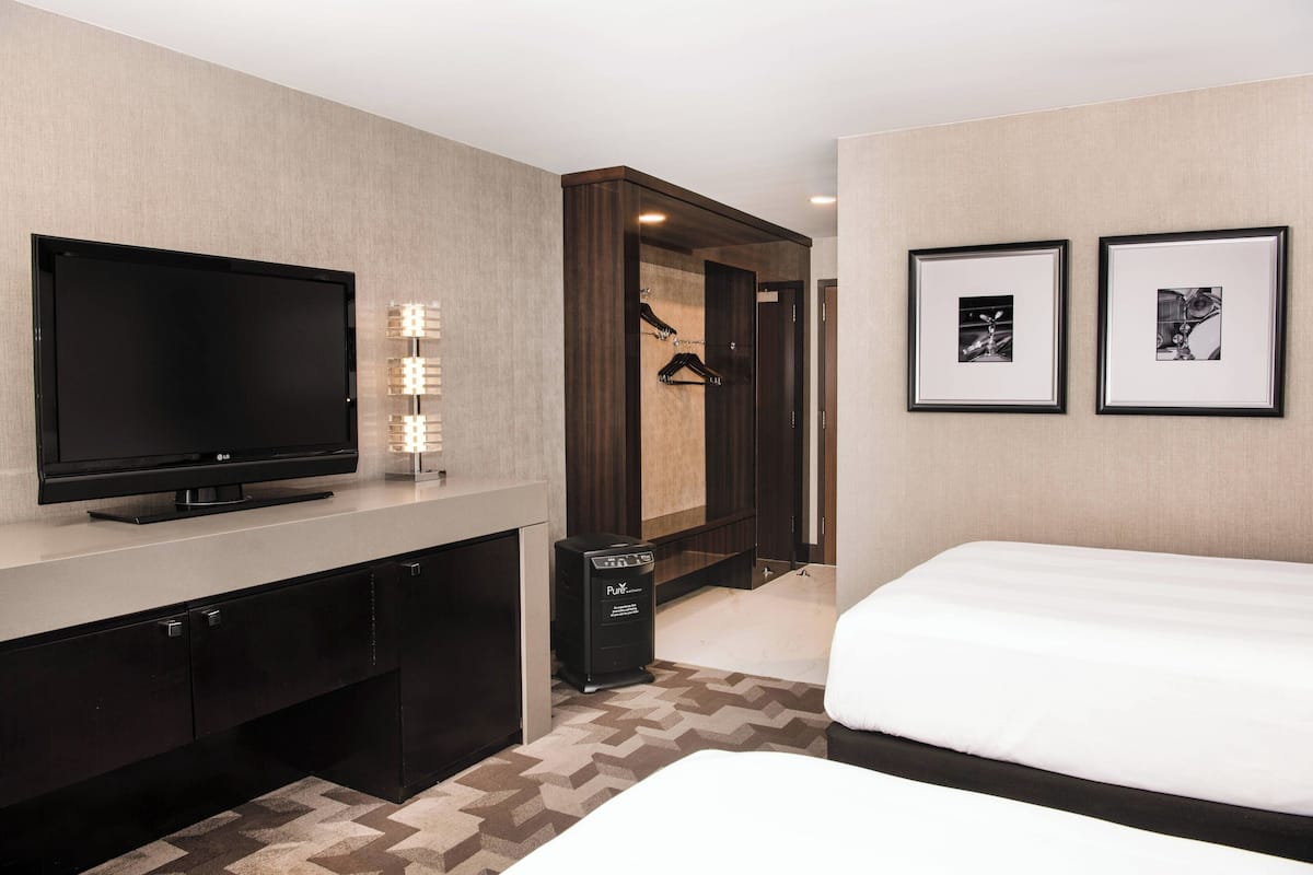 A hotel room with a flat-screen TV, a minibar, a bed, a mirror, and two framed pictures on the wall.
