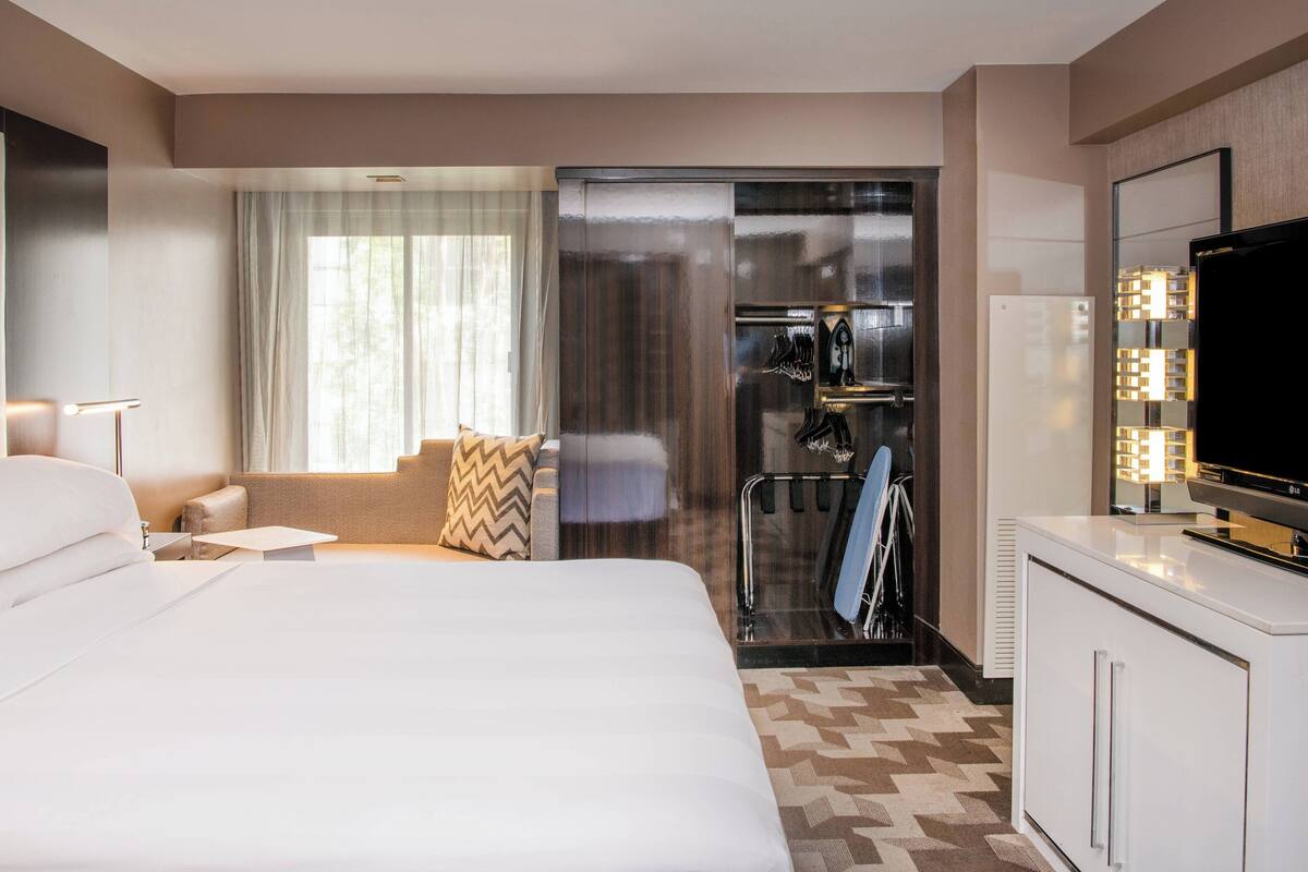 A hotel room with a large bed, a small table, a sofa, a TV, a refrigerator, and a glass door leading to a balcony.