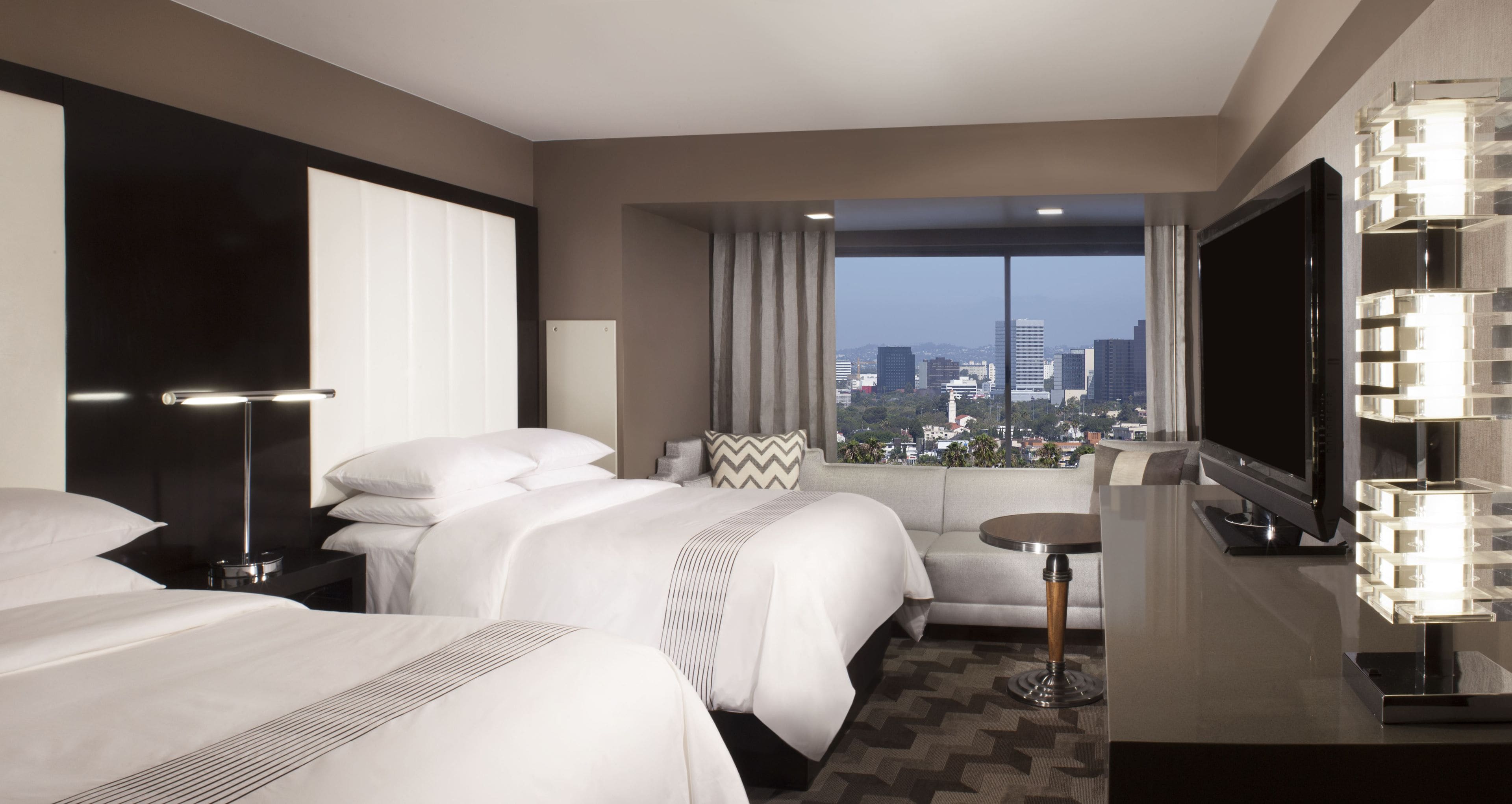 A modern hotel room with a large bed, a sofa, a small table, a TV, and a view of the cityscape.
