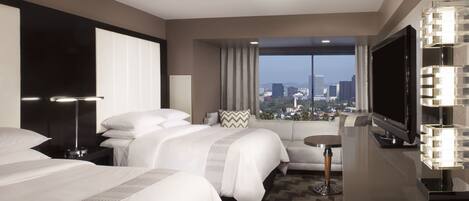 A modern hotel room with a large bed, a sofa, a small table, a TV, and a view of the cityscape.
