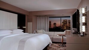 Quarto de hotel com uma cama grande, um sofá, uma escrivaninha e vista para o horizonte da cidade ao entardecer.