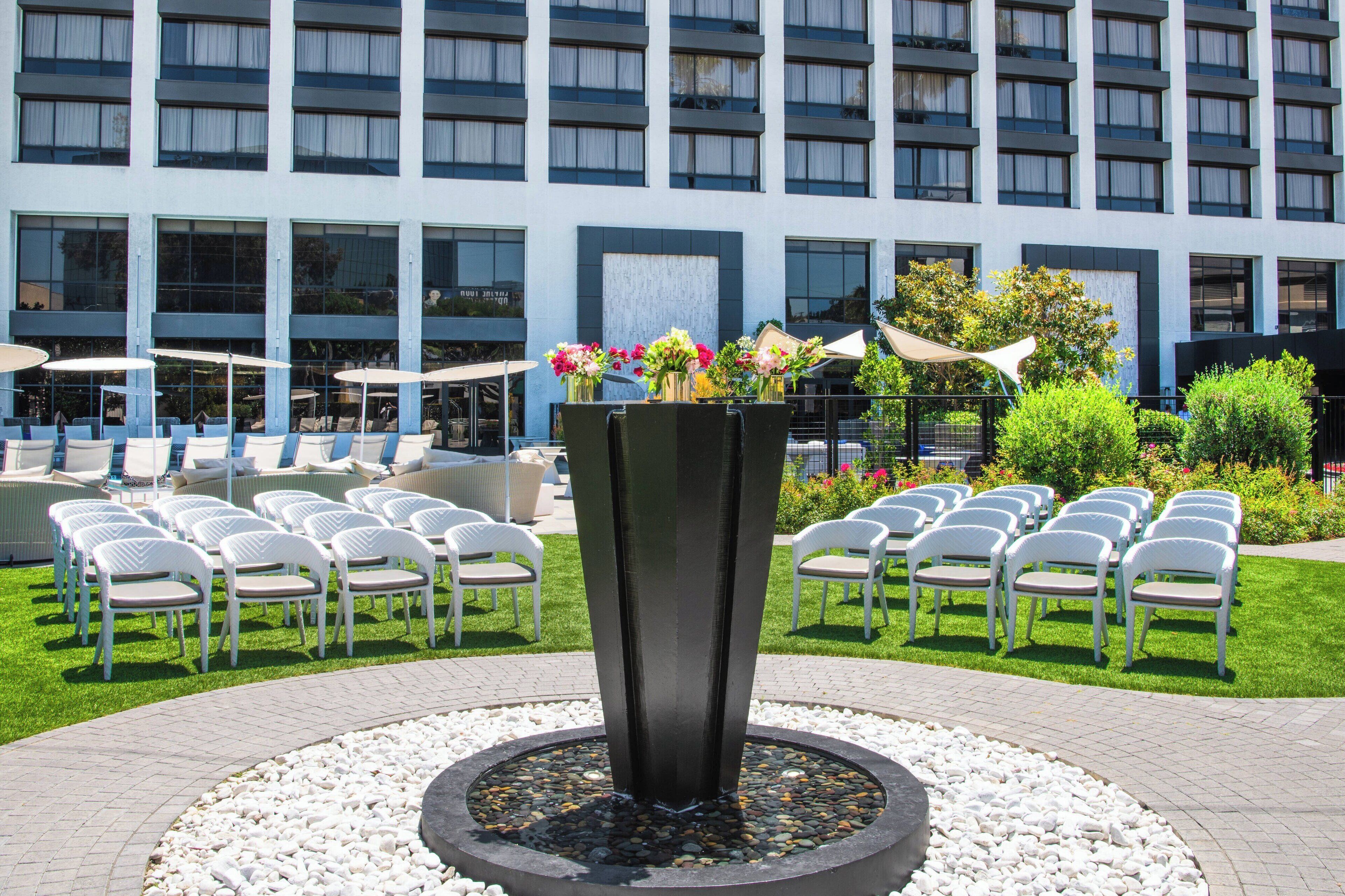 Un patio de hotel con una fuente central, mobiliario dispuesto y una fachada de edificio moderna al fondo.