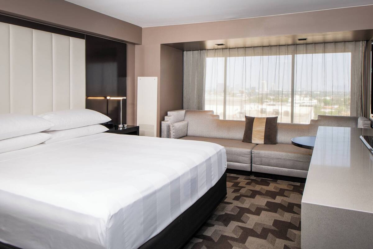 A hotel room with a large bed, a sofa, a small table, and a view of the city through large windows.