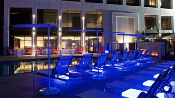 A poolside area with lounge chairs and umbrellas, a bar area with seating, and a building with large windows and a pool.