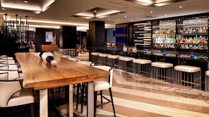 Uma área de bar moderna com uma longa mesa de madeira e banquetas brancas, uma estante de bebidas bem abastecida e luminárias contemporâneas.