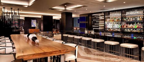 Un área de bar moderna con una larga mesa de madera y taburetes blancos, una estantería repleta de licores e iluminación contemporánea.