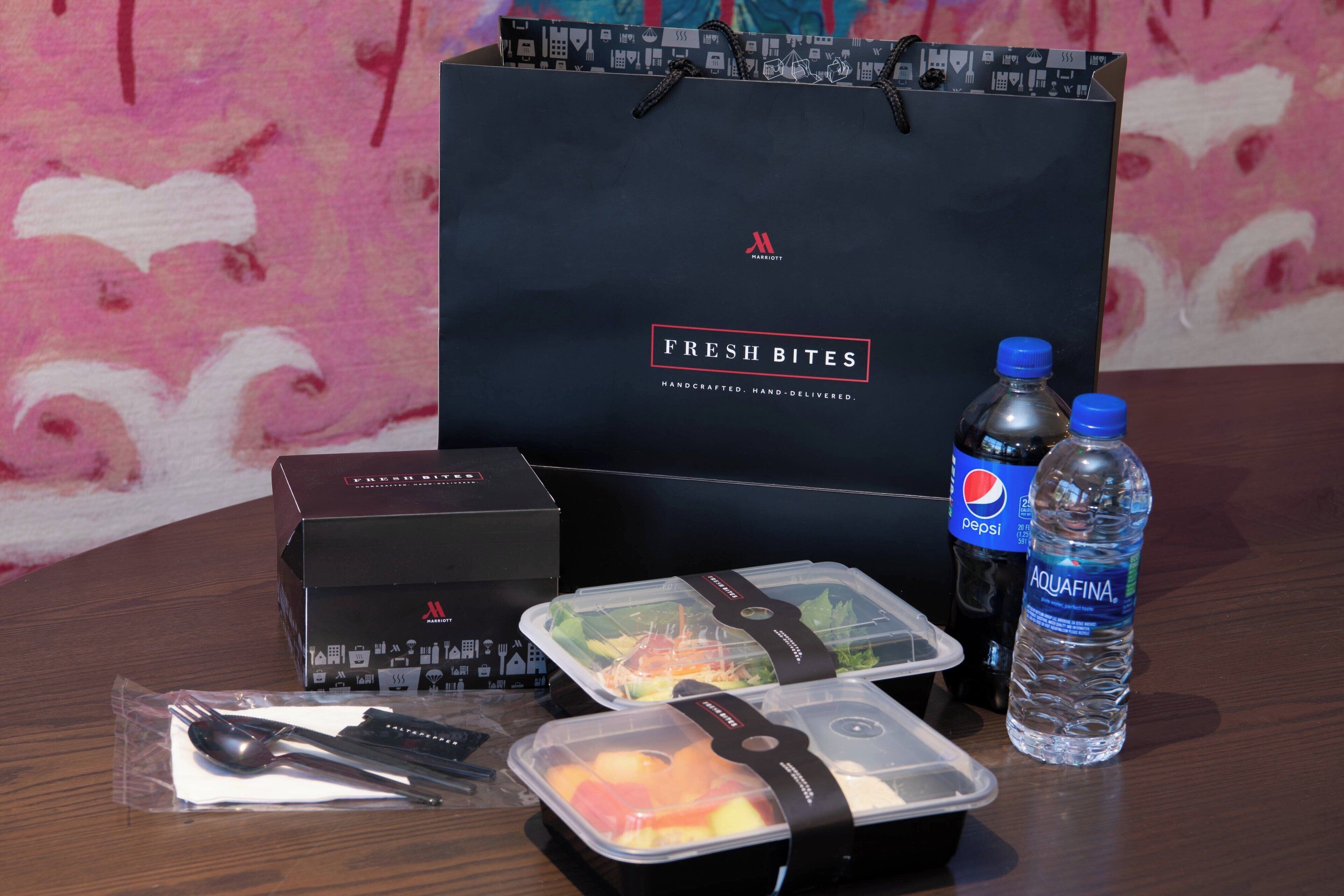 Een hotelkamer met een houten tafel, een zwarte Marriott tas, twee waterflessen, een Pepsi-fles en een maaltijdbox met bestek.