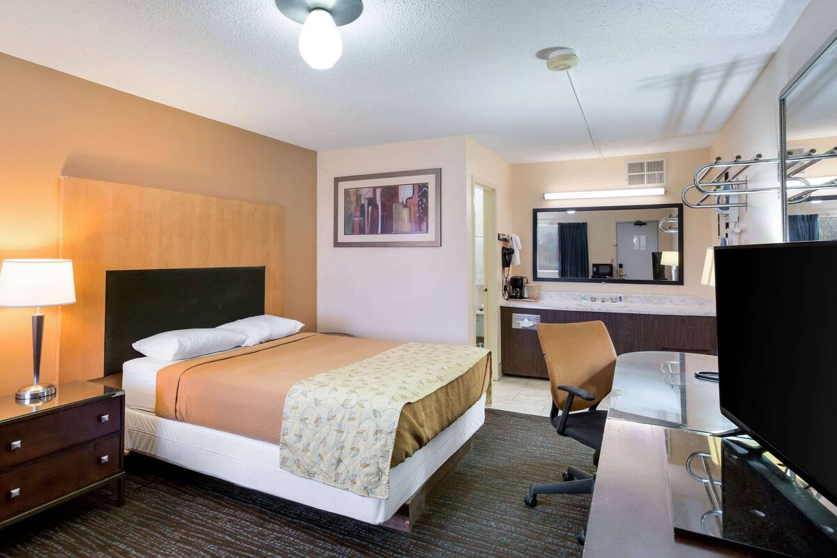 Una habitación de hotel con cama, escritorio, silla, televisor, lámpara, espejo y un cuadro en la pared.