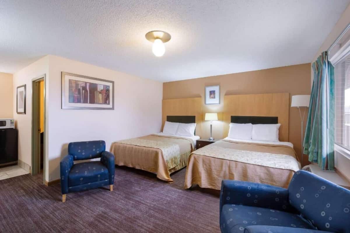 Une chambre d’hôtel avec deux lits, un bureau, une chaise, un tableau au mur et une fenêtre avec des rideaux.