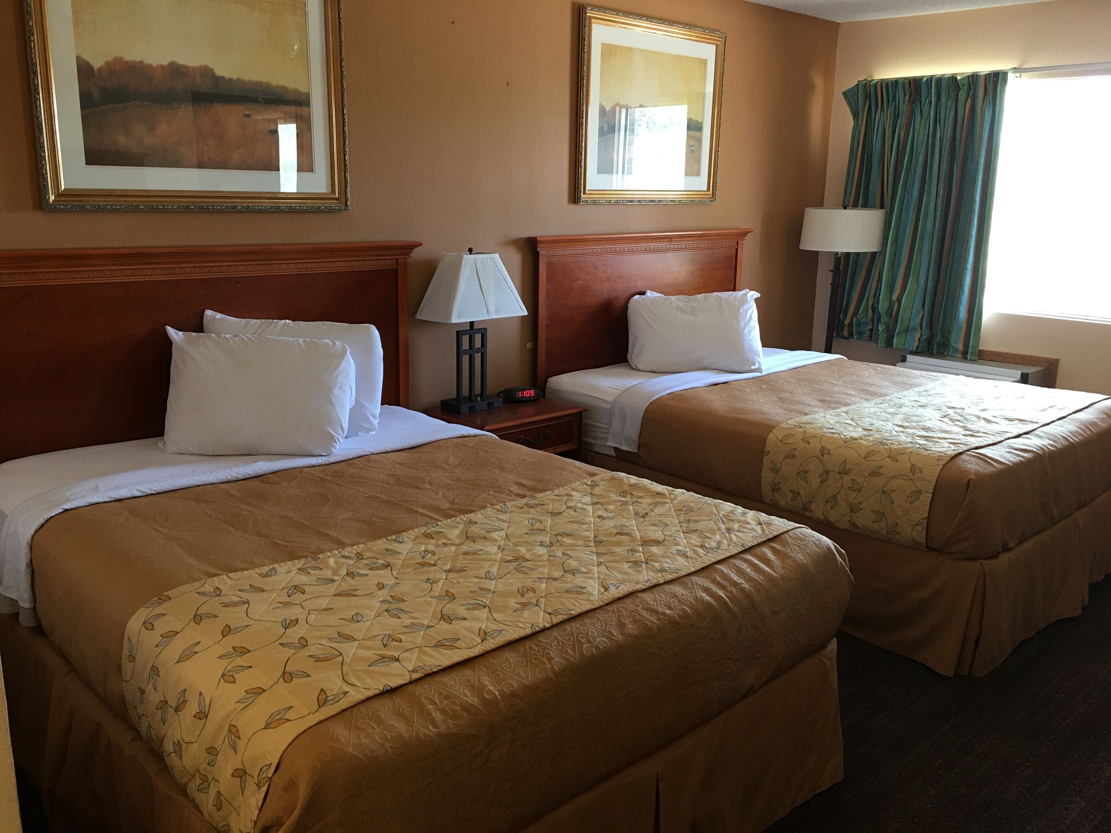A hotel room with two beds, each with a wooden headboard, white bedding, and a floral-patterned duvet. There are two bedside tables with lamps, a large framed painting above the left bed, and a window with curtains.