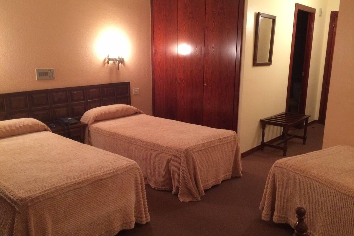 A hotel room with two single beds, a wooden wardrobe, a bedside table, a mirror, and a small bench.