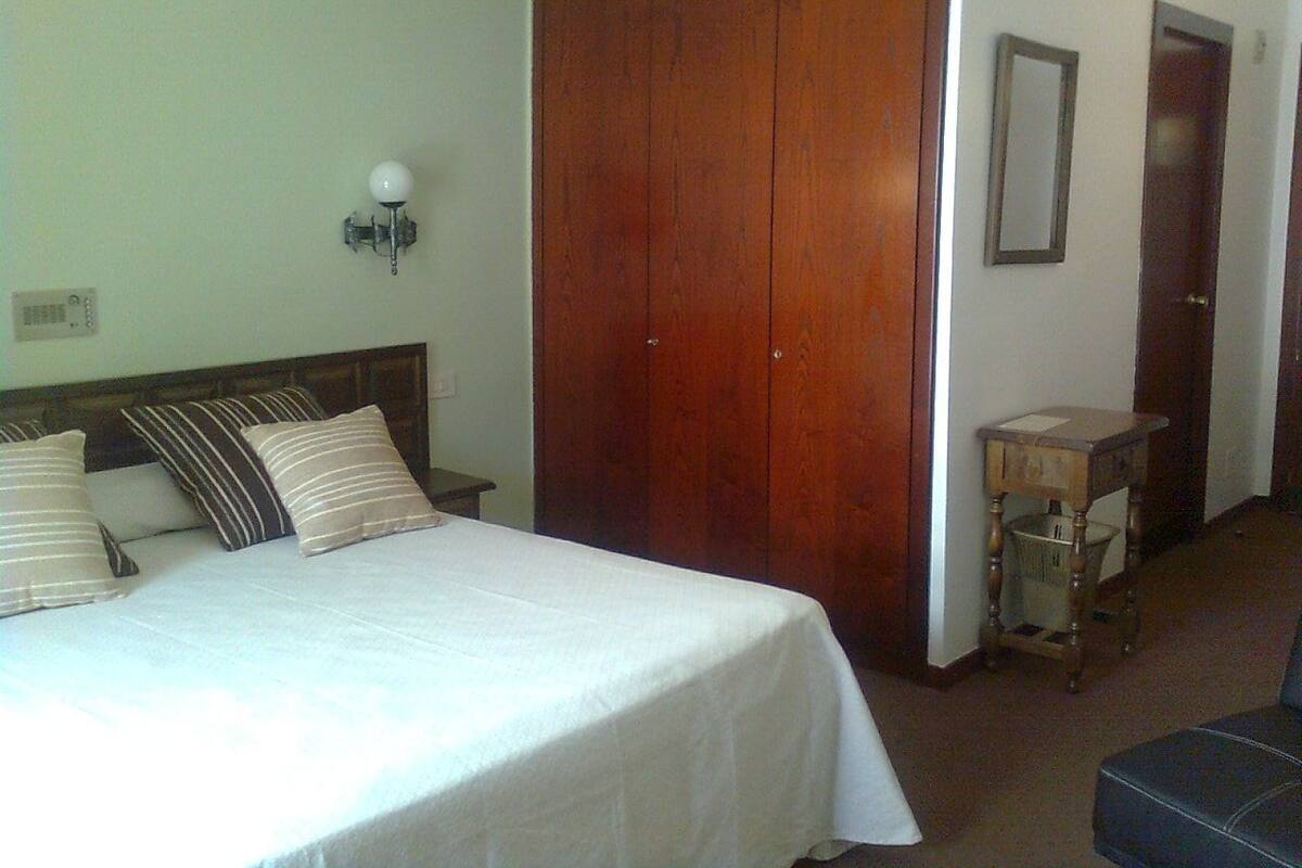 A hotel room with a bed, wooden wardrobe, bedside table, lamp, and a mirror on the wall.