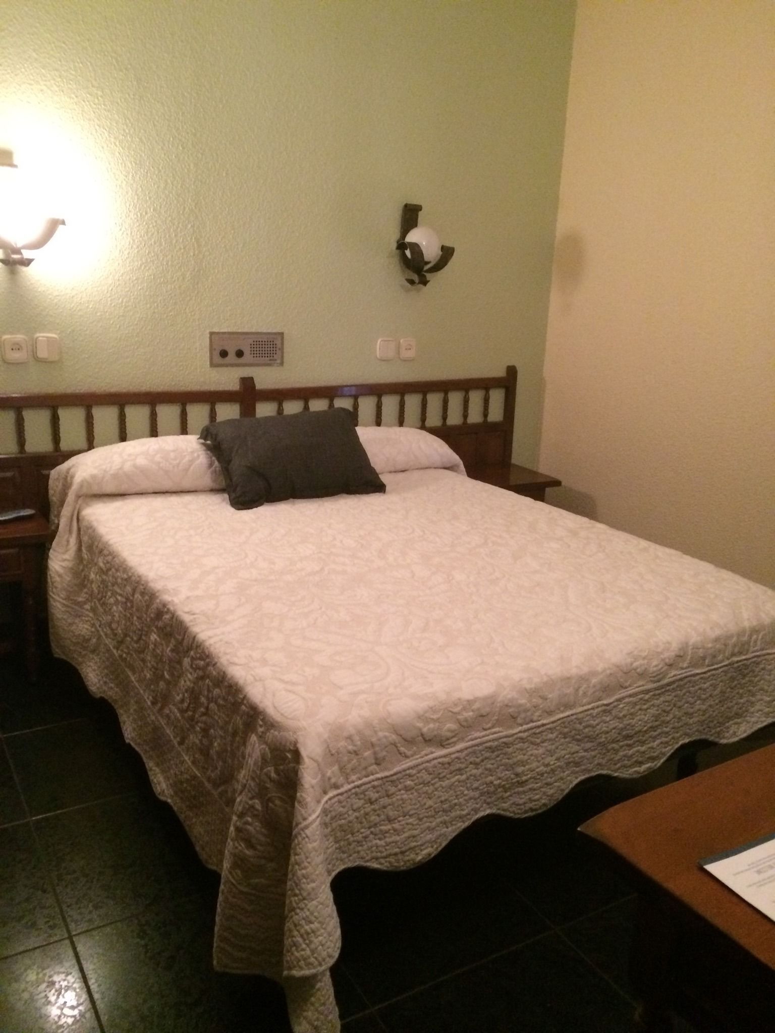 Een net opgemaakt bed met een witte dekbedovertrek en één donker kussen. Er is een houten hoofdbord, een klein nachtkastje en een wandlamp.
