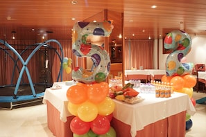 Quarto com um trampolim, uma mesa com balões e lanches, e uma grande decoração com um balão do número 2.