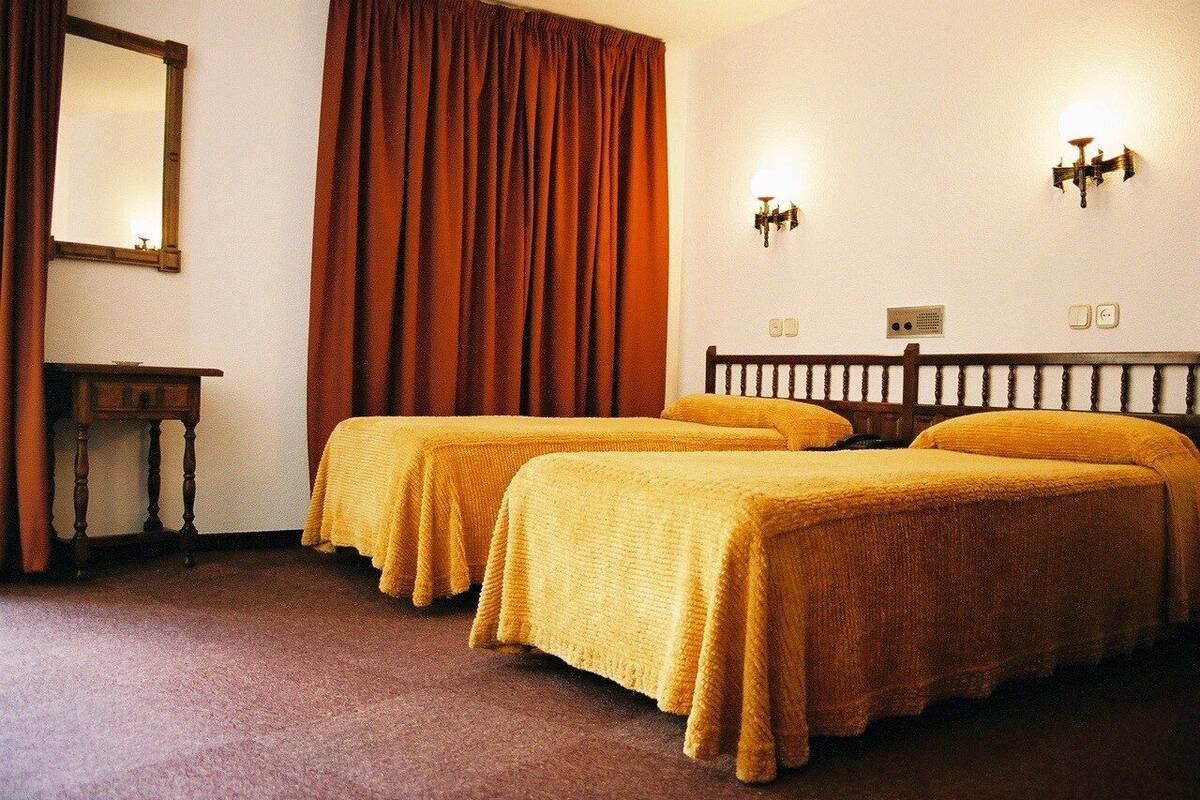 A hotel room with two beds, a wooden headboard, a bedside table, a mirror, and a large window with curtains.