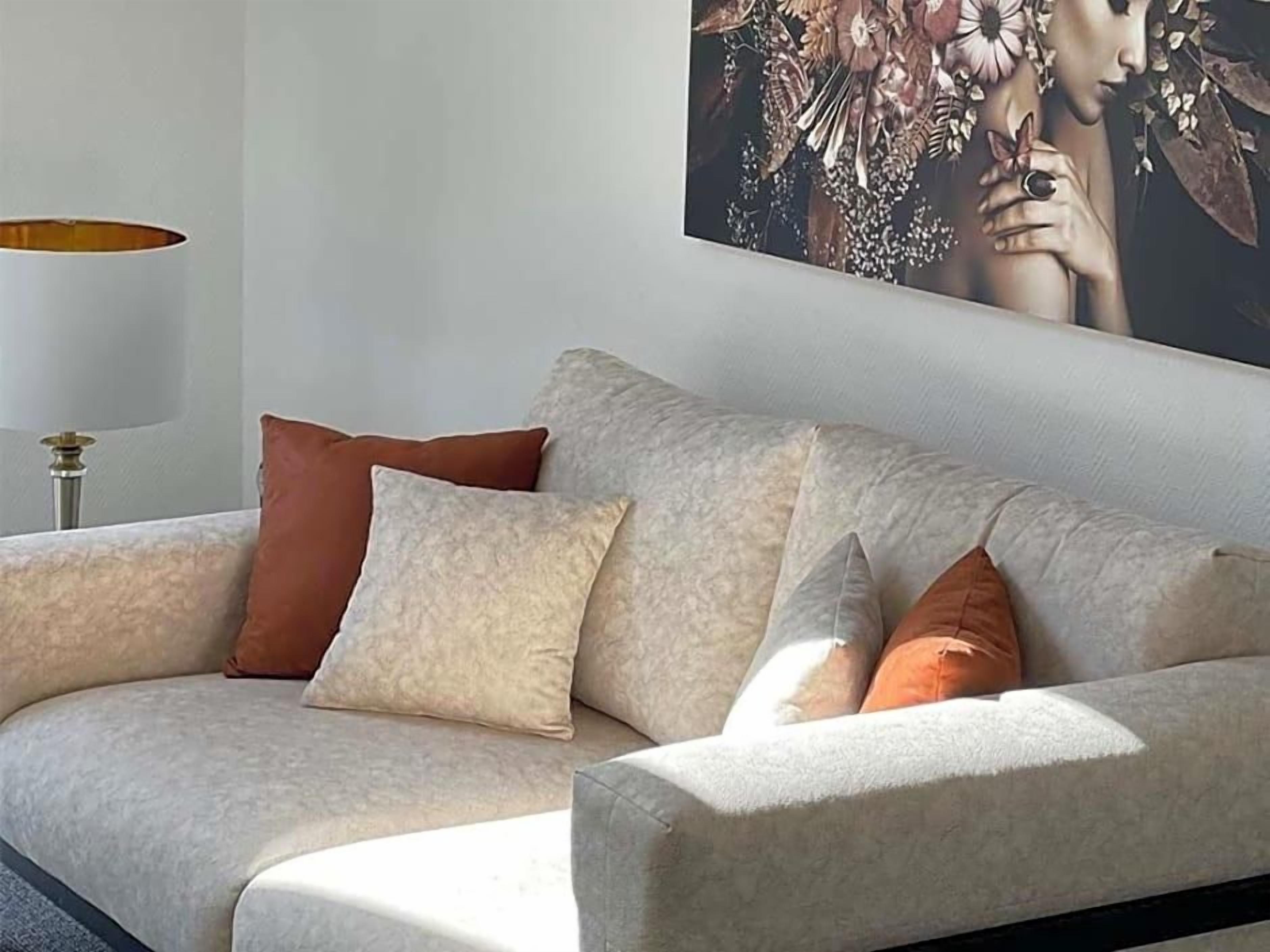Ein Sofa mit mehreren Kissen in einer neutralen Farbgebung, vor einer Wand mit einem gerahmten Kunstwerk.