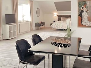 A modern hotel room with a bed, a TV, a dining area with chairs, a table, and a painting on the wall.