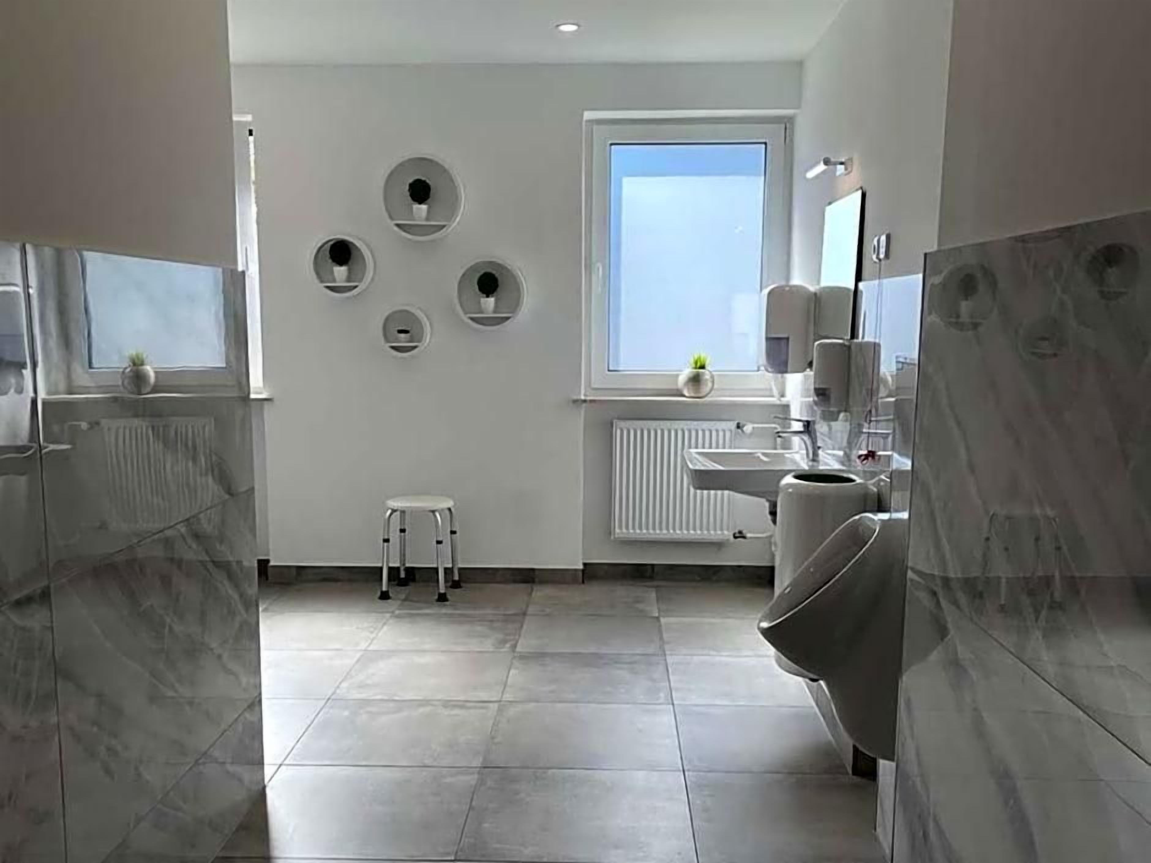Ein modernes Badezimmer mit Toilette, Waschbecken und einem kleinen Hocker. An den Wänden sind runde Dekorationen und ein Fenster für natürliches Licht vorhanden.