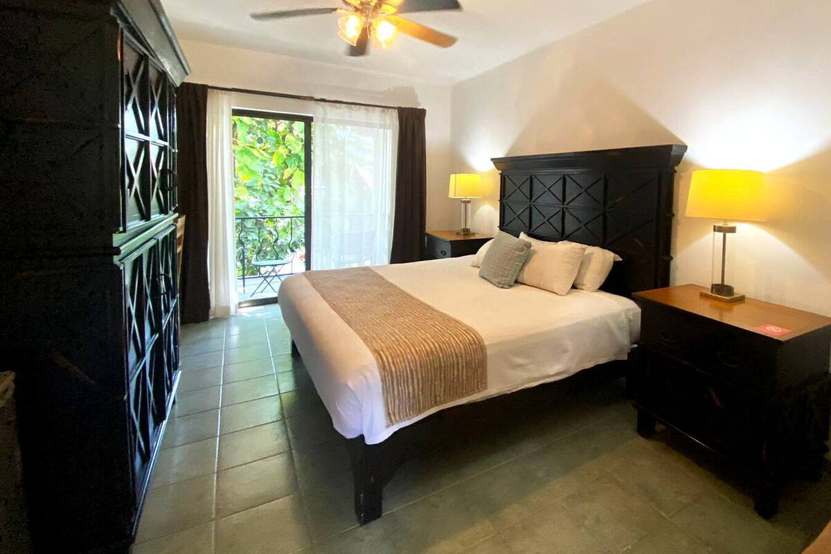 Une chambre d’hôtel avec un lit, deux tables de chevet avec des lampes, un ventilateur de plafond et une vue sur la verdure.
