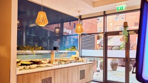 Un área de bufé con un expositor de vidrio, mostrador de madera, luces colgantes y vistas al exterior a través de amplias ventanas.