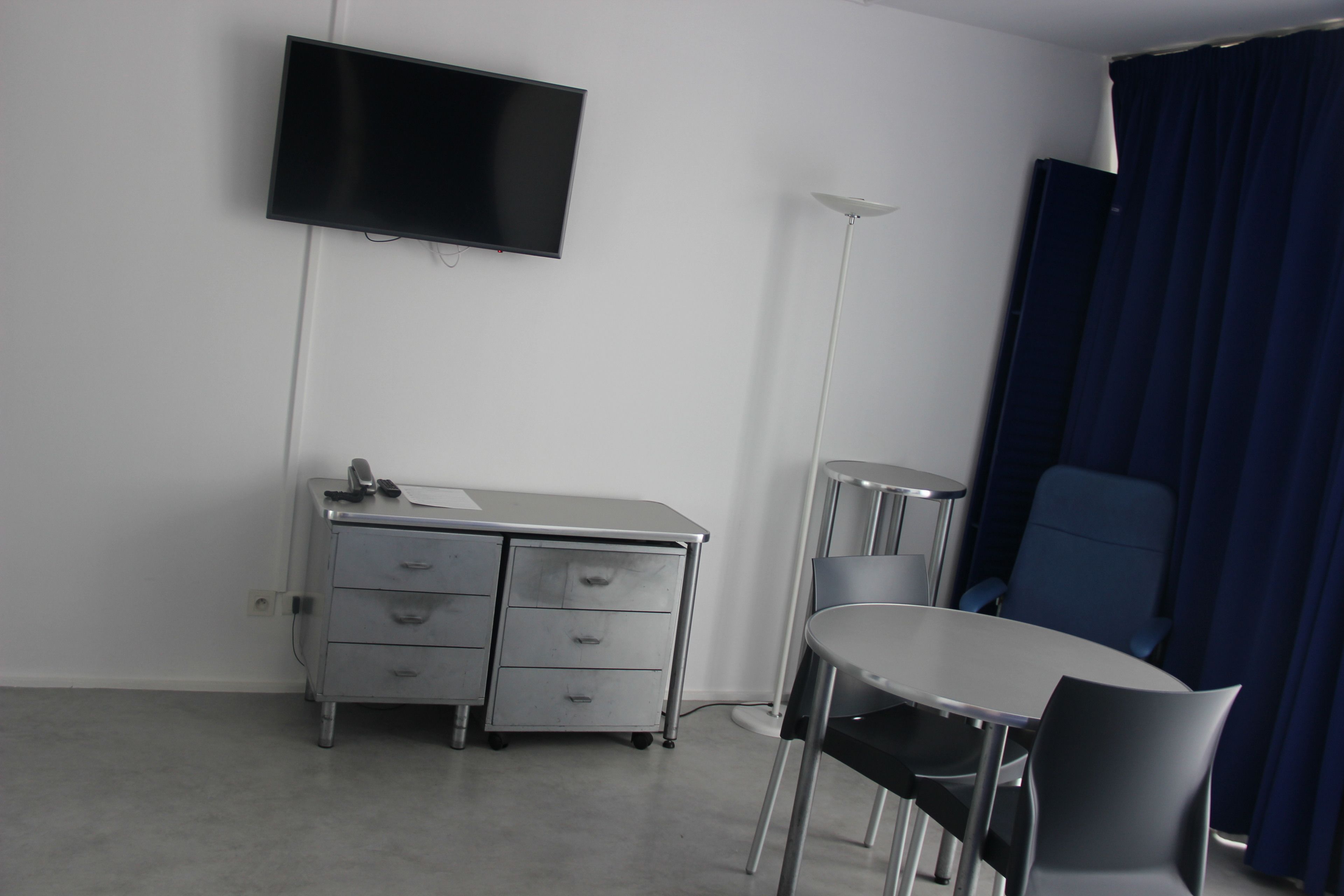 Ein Hotelzimmer mit Flachbildfernseher an der Wand, einem Schreibtisch mit Schubladen, einem Sessel und einer Stehlampe.