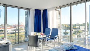 A modern hotel room with a large bed, a desk, two chairs, and a view of the city through large windows.