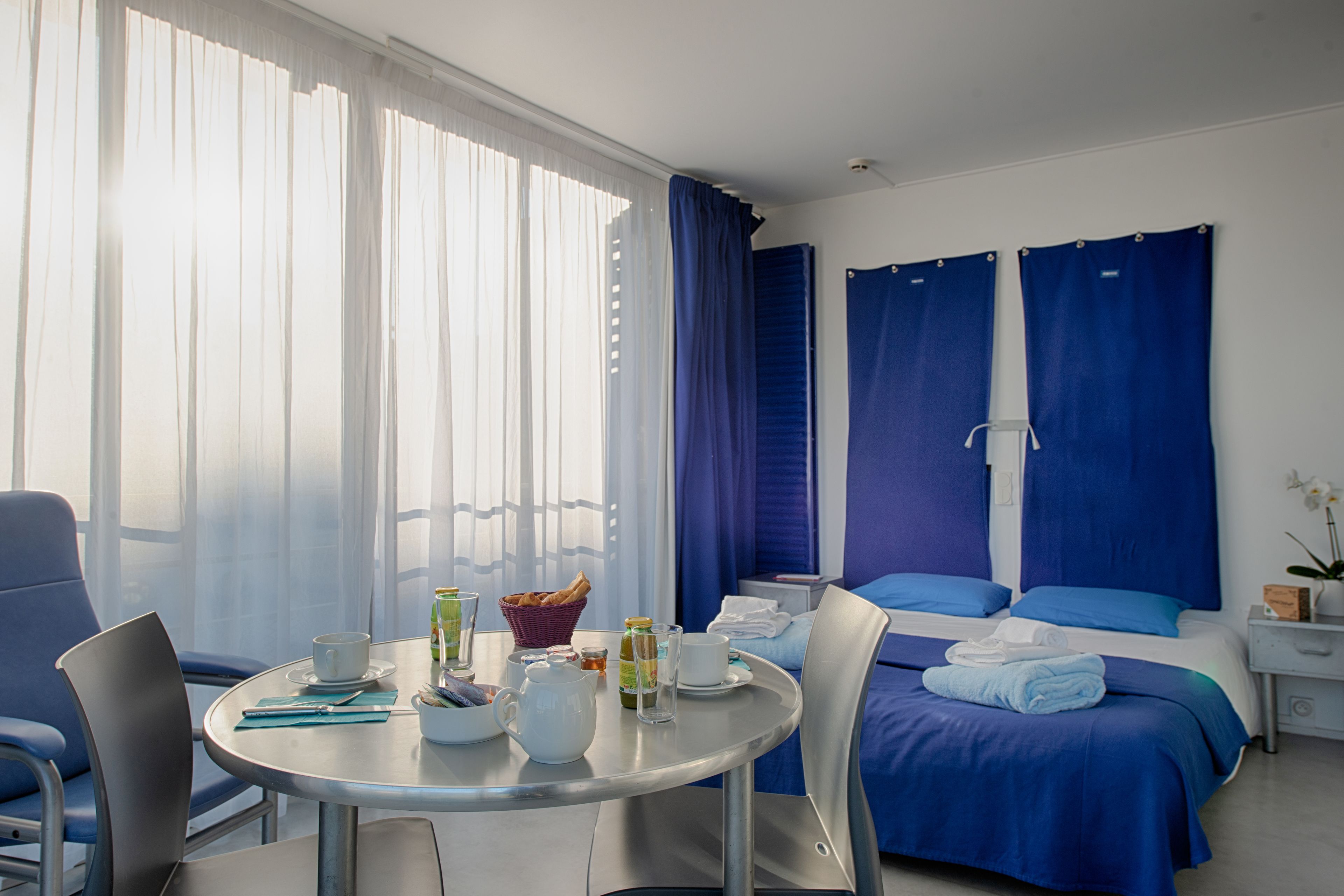A hotel room with a bed, a table with breakfast items, a chair, and a window with curtains.
