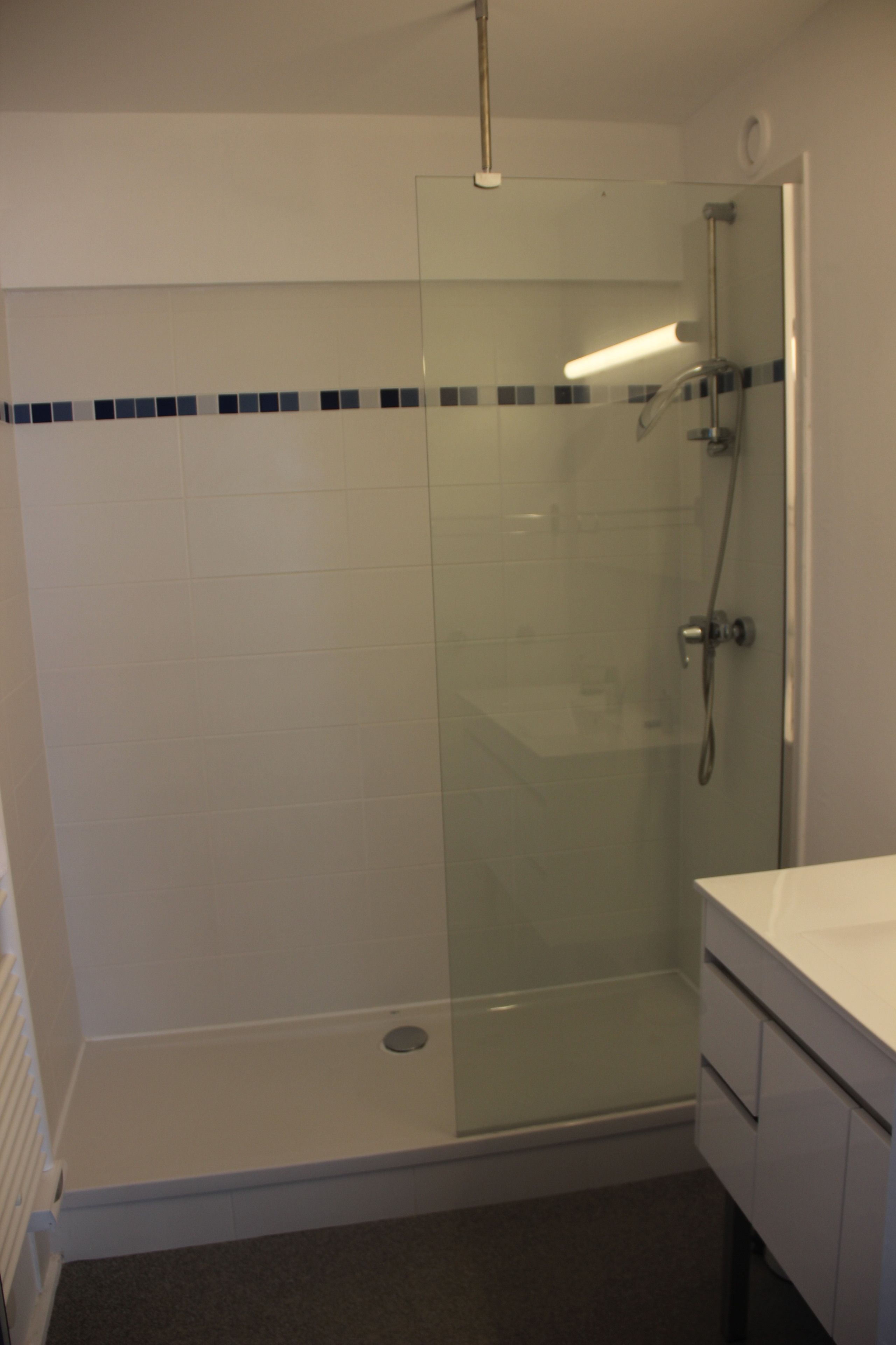 A glass-enclosed shower stall with a white tiled wall and a white countertop to the right.