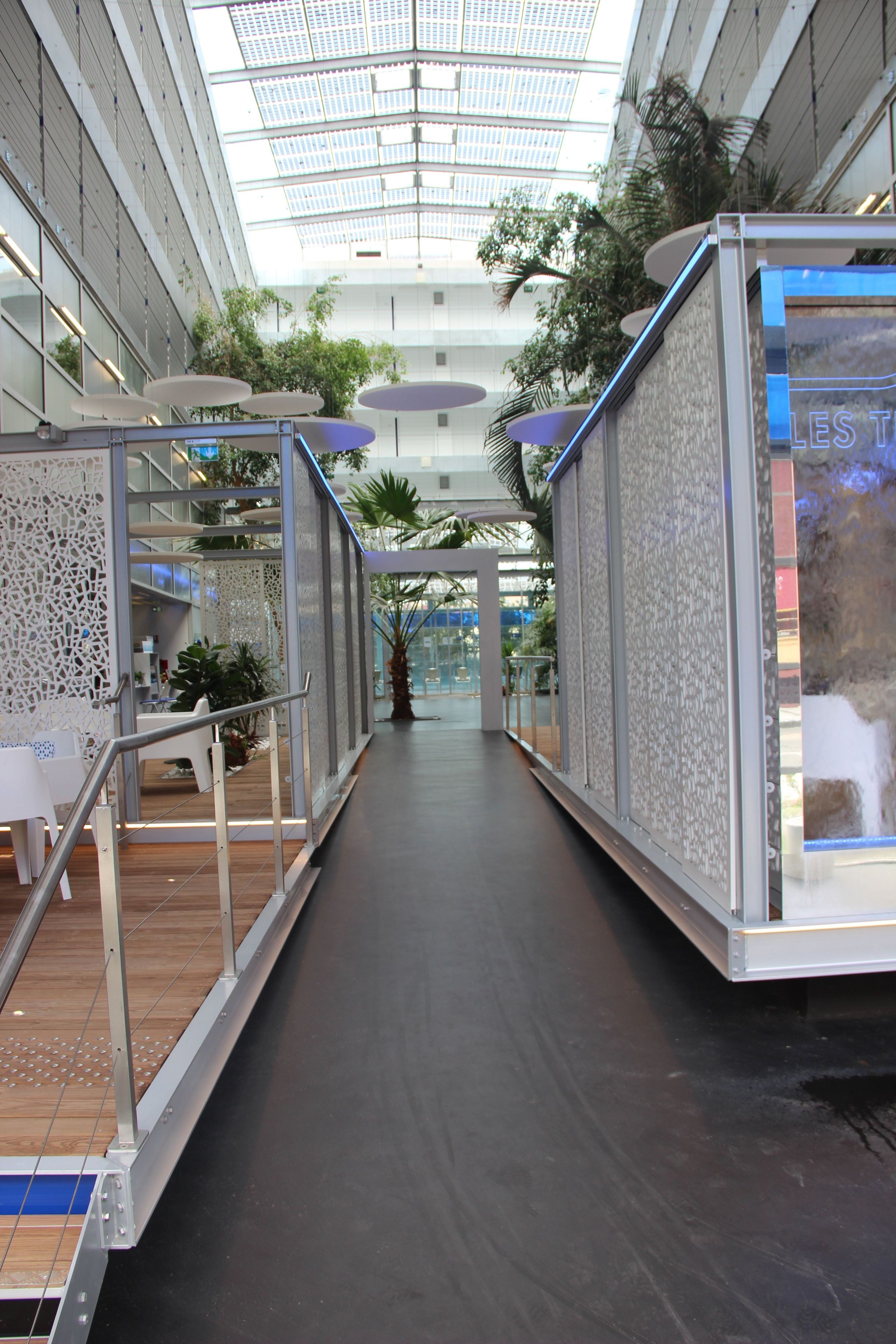 A modern indoor corridor with glass railings, wooden flooring, and potted plants.