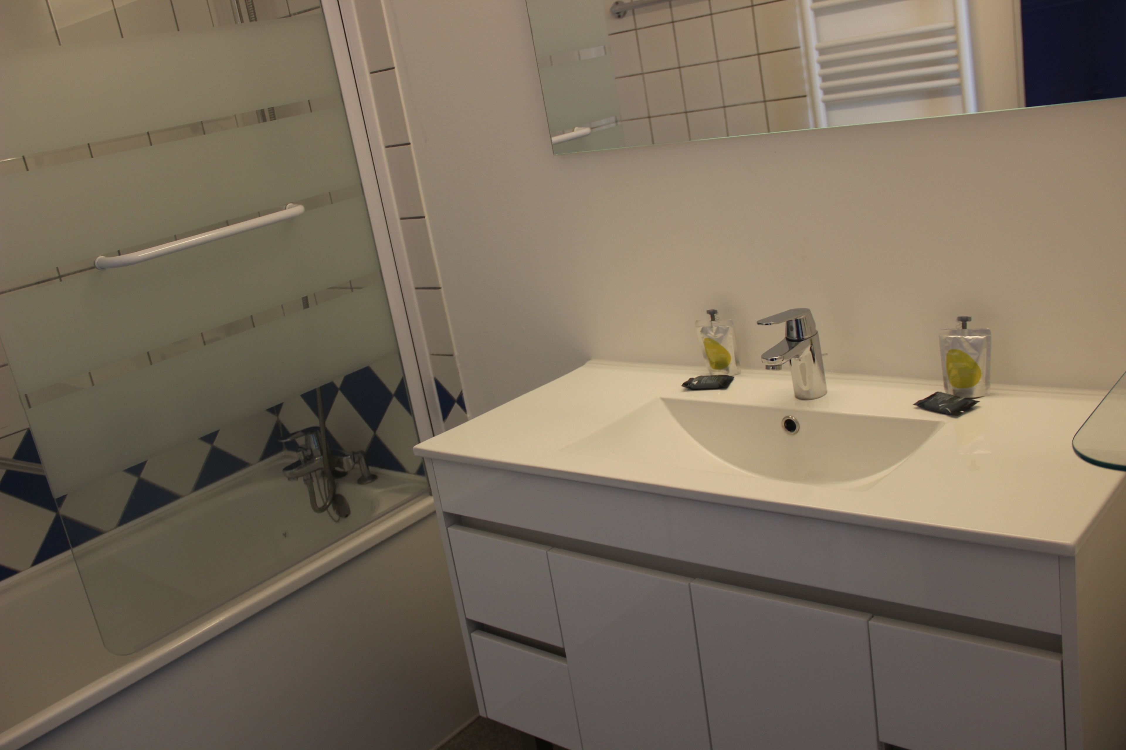 Une salle de bain avec un lavabo blanc, deux distributeurs de savon, un miroir et une cabine de douche en verre.