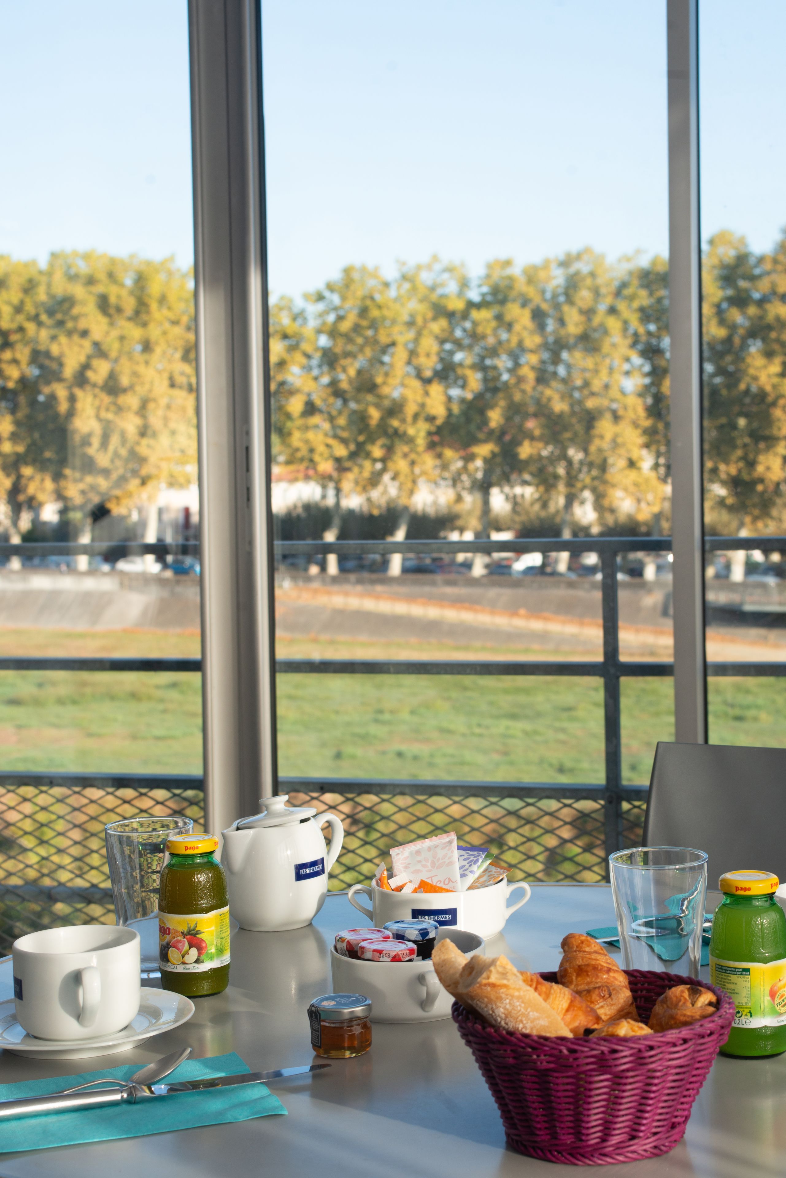 A breakfast table with a basket of croissants, a teapot, cups, and a bottle of juice.