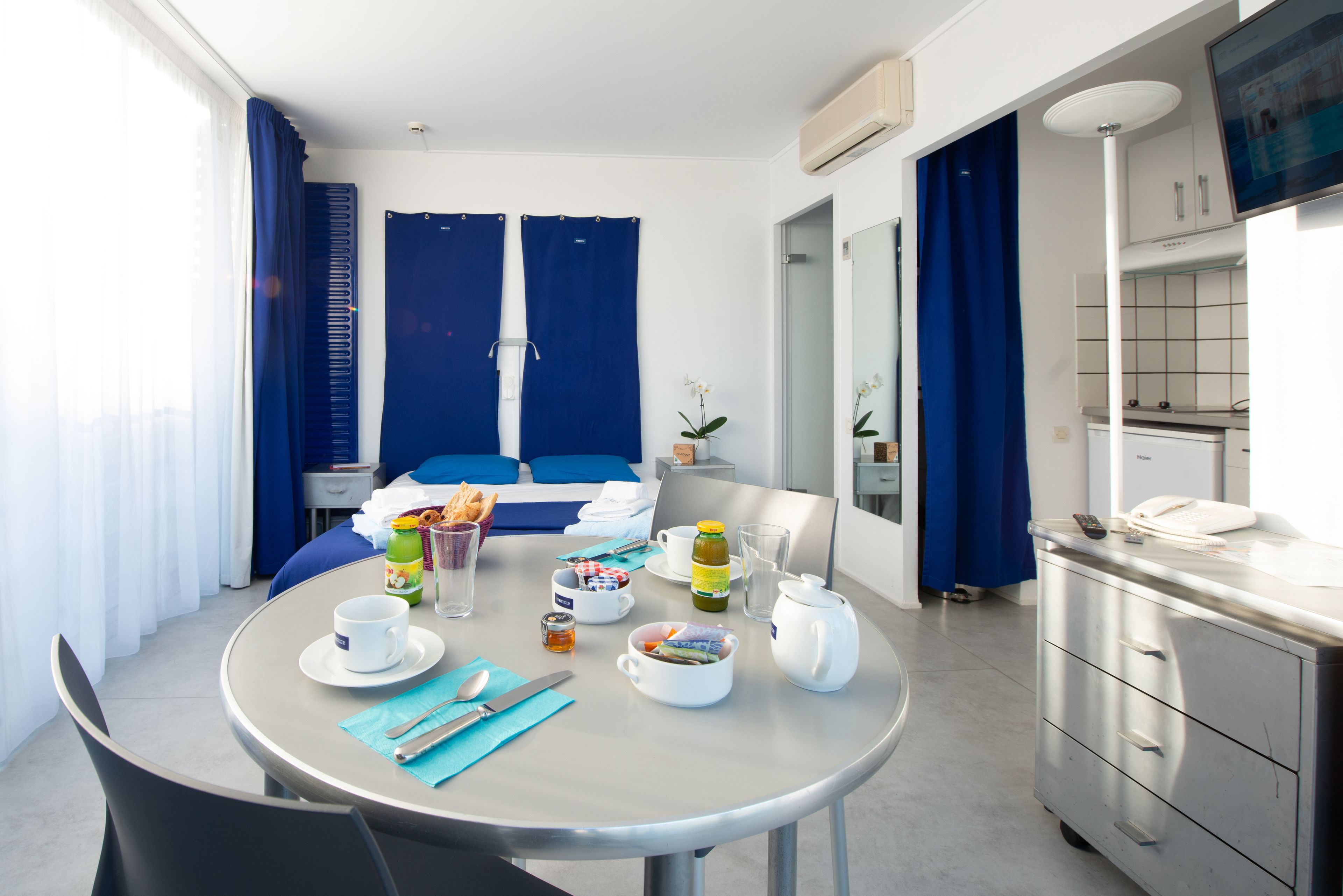 Une chambre d’hôtel avec une table à manger dressée pour le petit-déjeuner, un lit avec des rideaux bleus, un petit coin cuisine et une salle de bain visible à travers une porte ouverte.