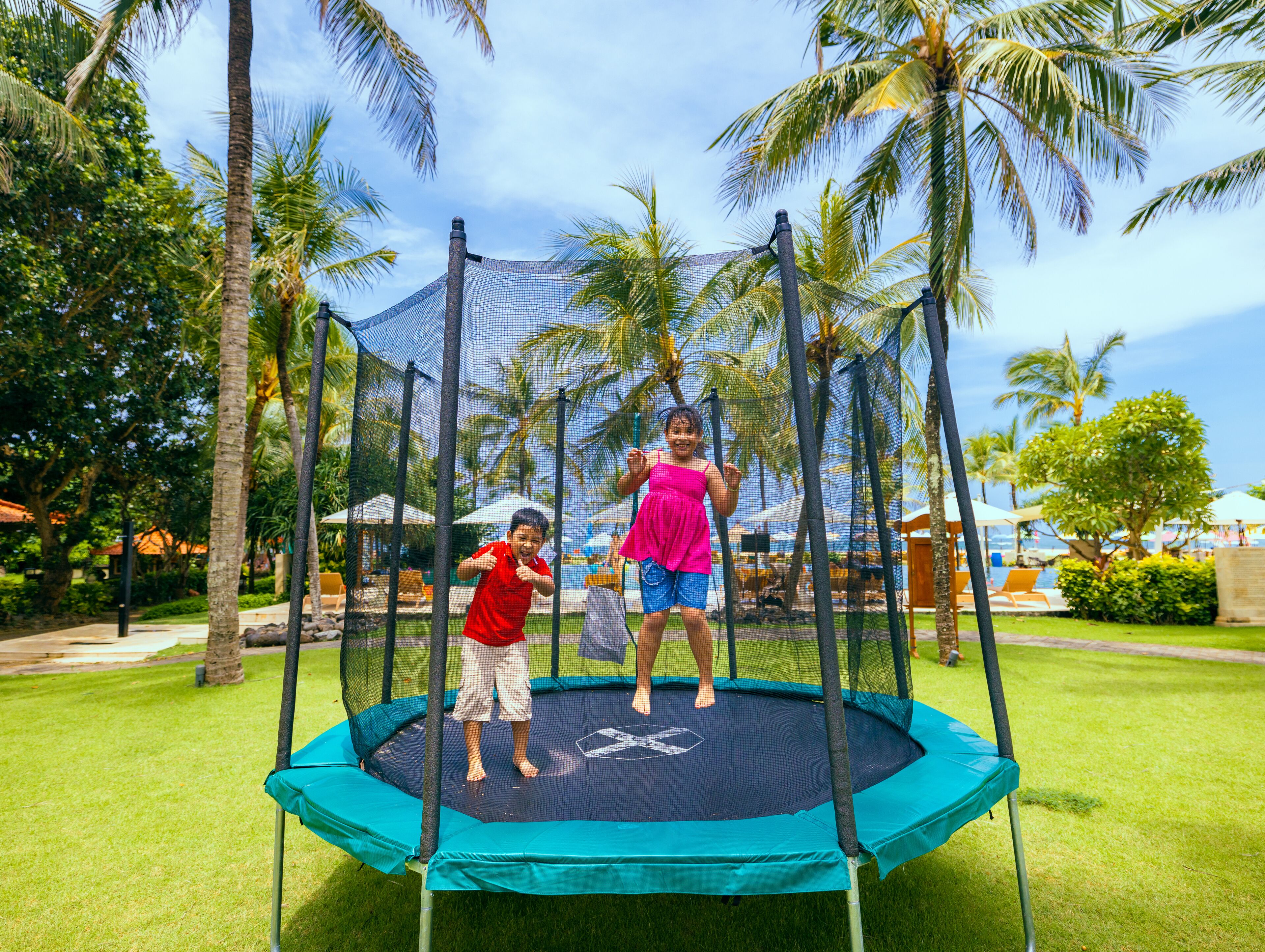 A trampoline in a grassy area with palm trees and umbrellas in the background.
