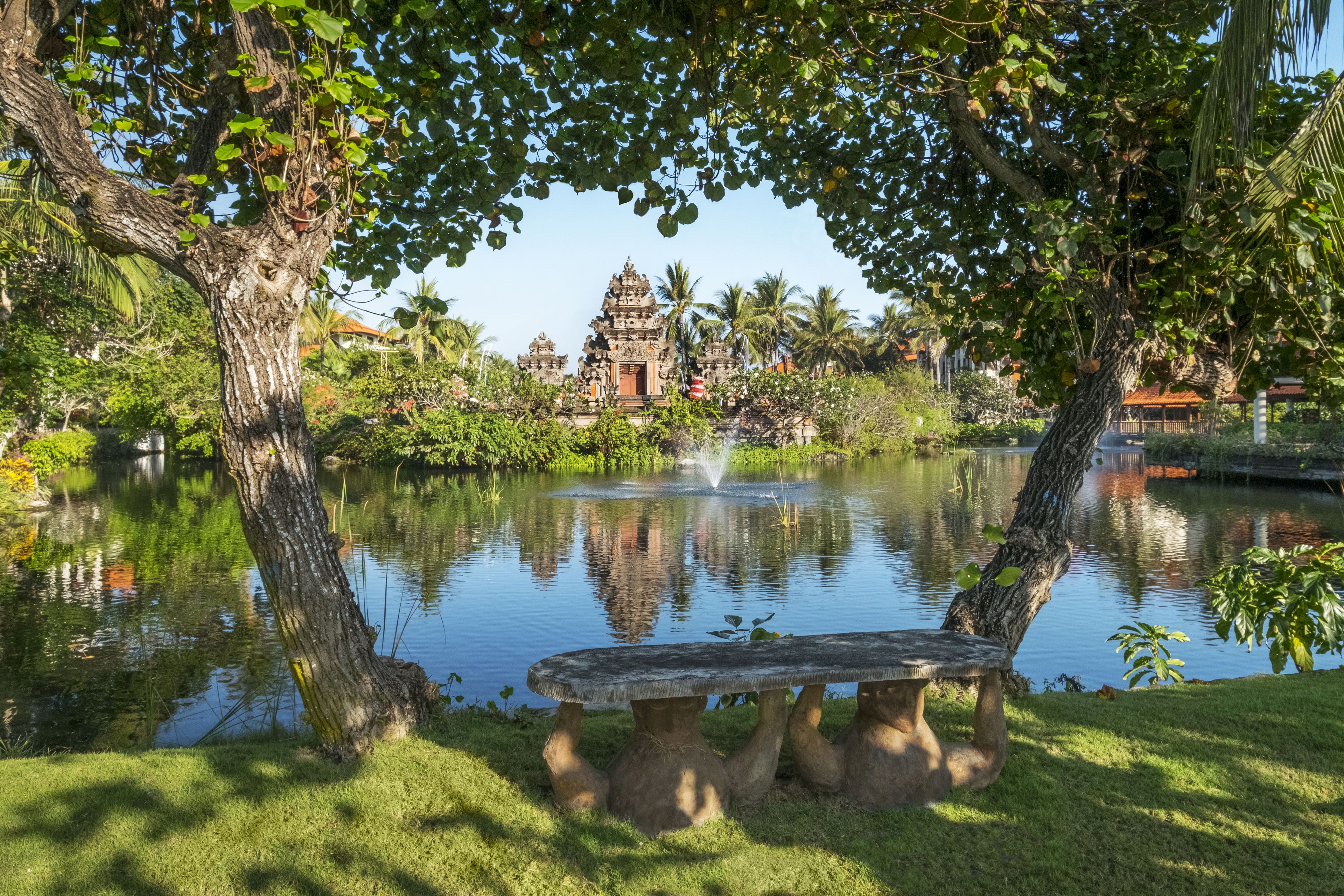 Um lago tranquilo com um banco de pedra, rodeado por vegetação exuberante e um templo ao fundo.