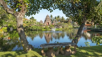 A serene pond with a stone bench, surrounded by lush greenery and a temple in the background.