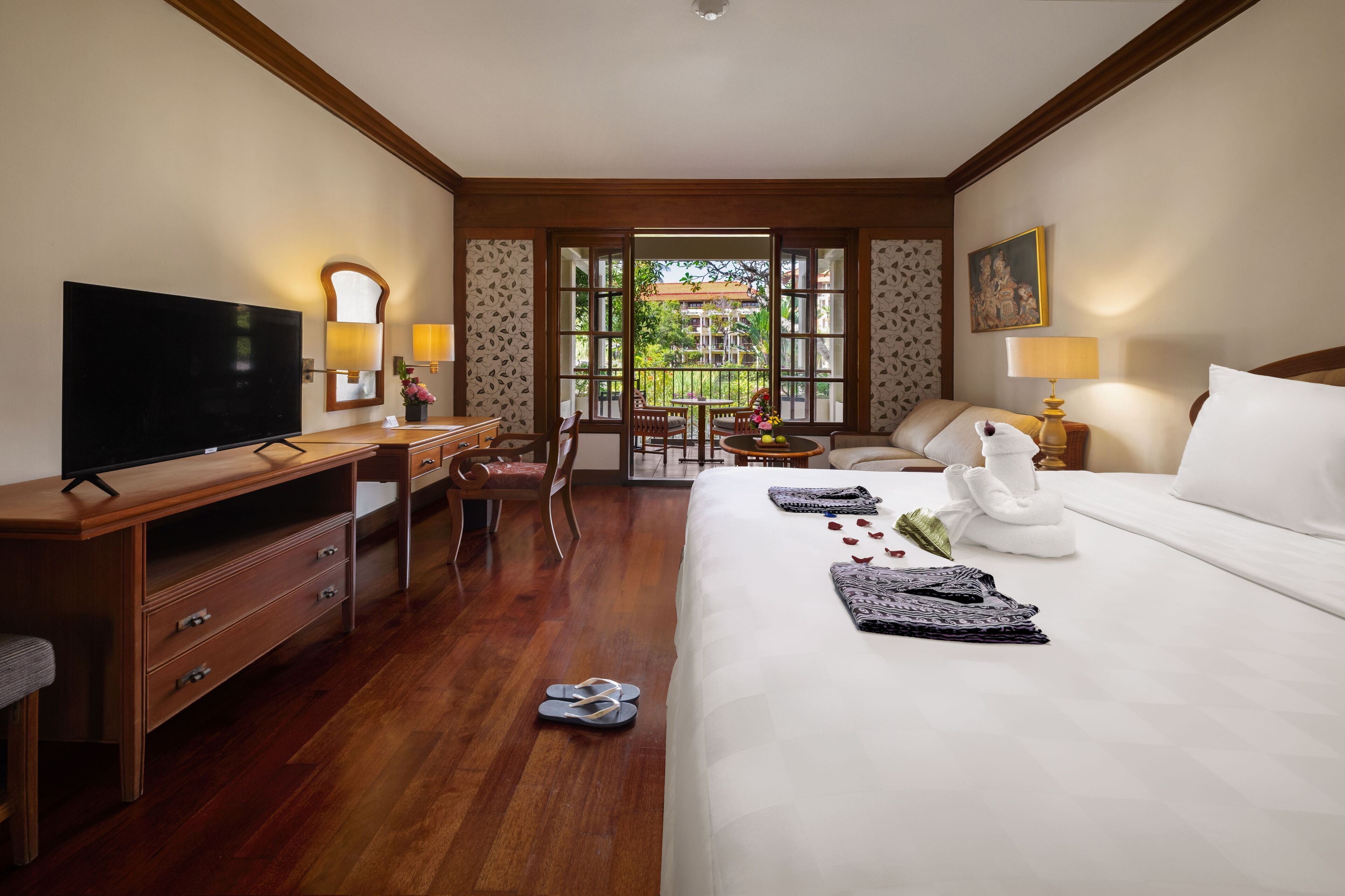 A hotel room with a bed, a TV, a desk, a chair, and a balcony with a view of greenery.