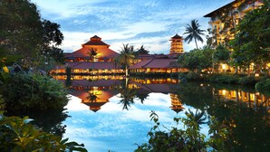 A traditional building with a reflection in the water, surrounded by greenery and palm trees.