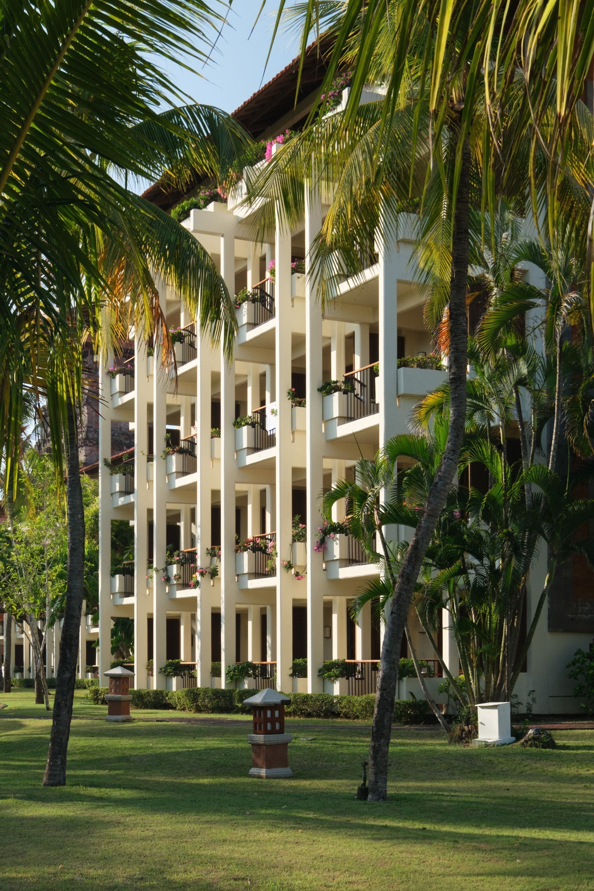 A multi-story hotel with balconies, palm trees, and a well-maintained lawn.