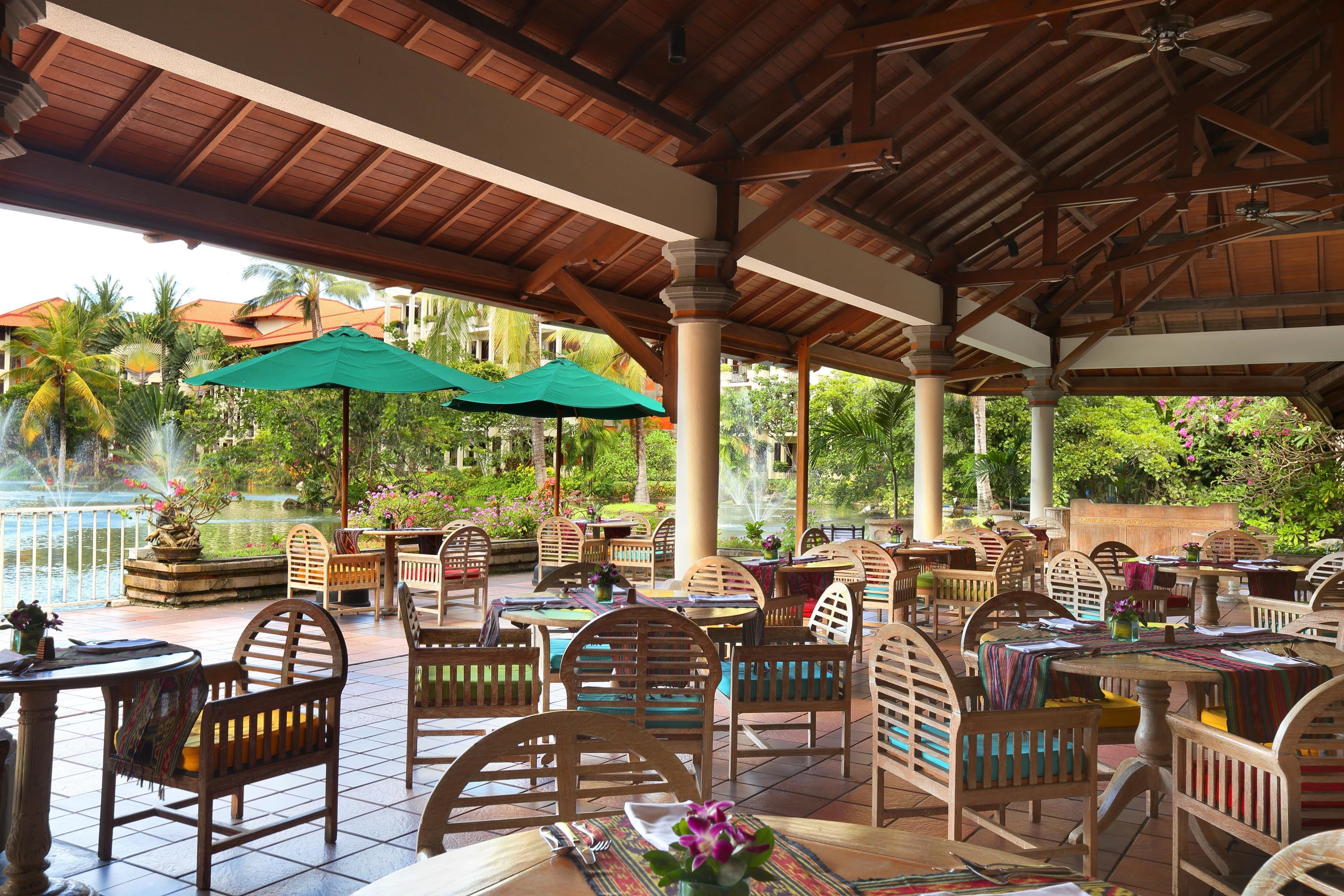 An outdoor patio with wooden furniture, green umbrellas, and a view of a garden and buildings.