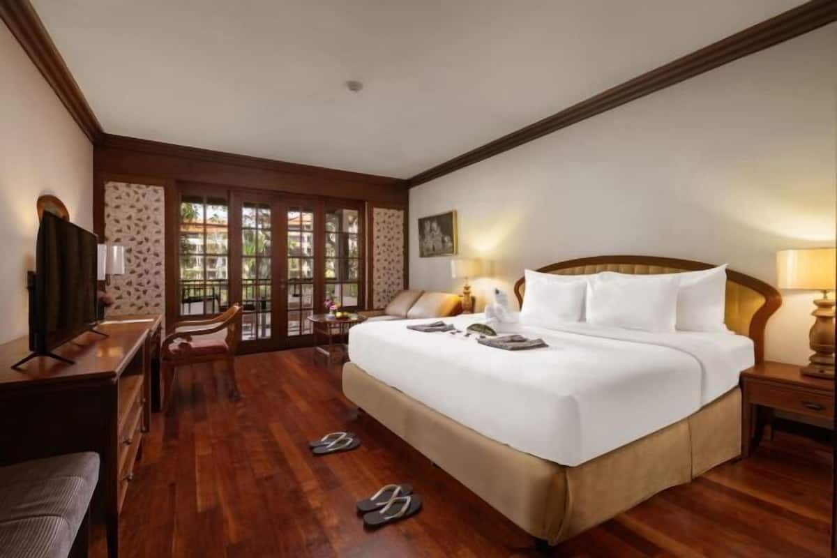 A hotel room with a large bed, a desk, a chair, a TV, and a window with a view of greenery.